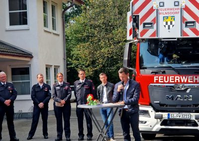 Landrat Müller steht vor einem Feuerwehrfahrzeug und hält ein Grußwort. Im Hintergrund stehen einige Feuerwehrleute und Wilnsdorfs Bürgermeister Gieseler.
