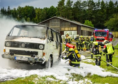 Mehrere Feuerwehrleute löschen das brennende Fahrzeug mit Schaum.