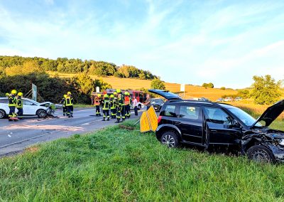 Auf der L893 steht eines der beiden Unfallfahrzeuge. Feuerwehrleute kehren Trümmerteile zusammen.