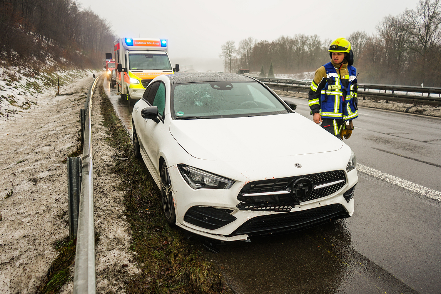 Verunfallter weißer Pkw steht auf dem Seitenstreifen einer nassen Autobahn; die Front und die Windschutzscheibe sind beschädigt. Daneben ein Feuerwehrmann in Einsatzkleidung, im Hintergrund ein Rettungswagen mit eingeschaltetem Blaulicht bei winterlichen Straßenverhältnissen.