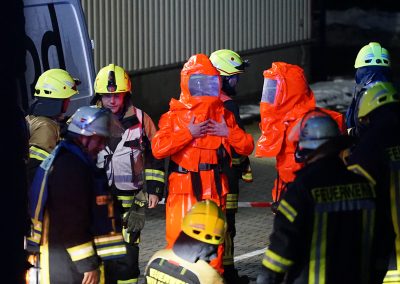 Mehrere Feuerwehrkräfte stehen auf dem Betriebsgelände zusammen; zwei Einsatzkräfte tragen orangefarbene Chemikalienschutzanzüge.