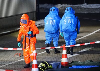Drei Einsatzkräfte in Chemikalienschutzanzügen in einem abgesperrten Bereich; eine Person im orangefarbenen Schutzanzug geht im Vordergrund, zwei weitere in blauen Anzügen entfernen sich im Hintergrund auf dem Betriebsgelände.