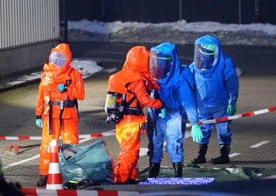 Vier Einsatzkräfte in orange- und blaufarbenen Chemikalienschutzanzügen stehen in einem abgesperrten Einsatzbereich.