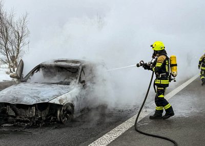 Eine Einsatzkraft löscht auf dem Standstreifen der Autobahn einen ausgebrannten Pkw; dichter Rauch steigt auf.