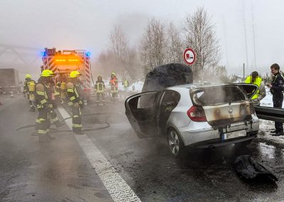 Mehrere Einsatzkräfte stehen in der Nähe des ausgebrannten und bereits gelöschten PKWs, der auf dem Standstreifen der Autobahn steht; leichter Rauch steigt auf.