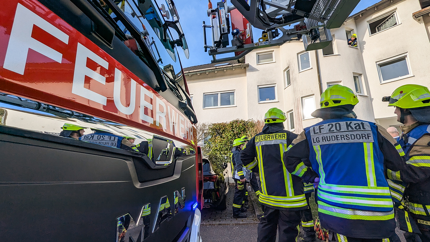 Nahaufnahme der Front eines Feuerwehrwagens. Rechts daneben stehen mehrere Einsatzkräfte der Feuerwehr vor einem Wohngebäude, über dem ein Teil einer Drehleiter zu sehen ist. Aus einem der oberen Fenster schaut ein Feuerwehrmann.