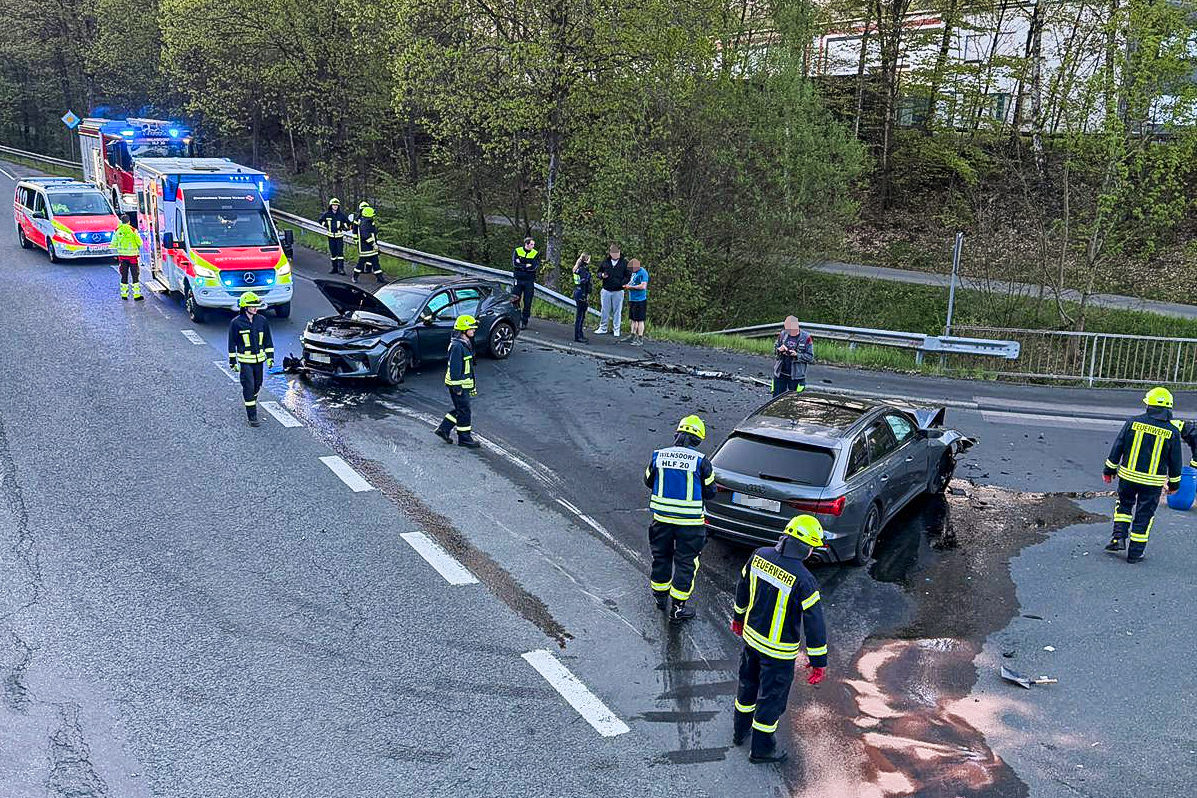 Die beiden beschädigten Pkw stehen auf der Fahrbahn, einer mit geöffneter Motorhaube, der andere mit stark deformierter Front. Feuerwehr und Rettungsdienst sind vor Ort, Einsatzkräfte kümmern sich um ausgelaufene Flüssigkeiten.