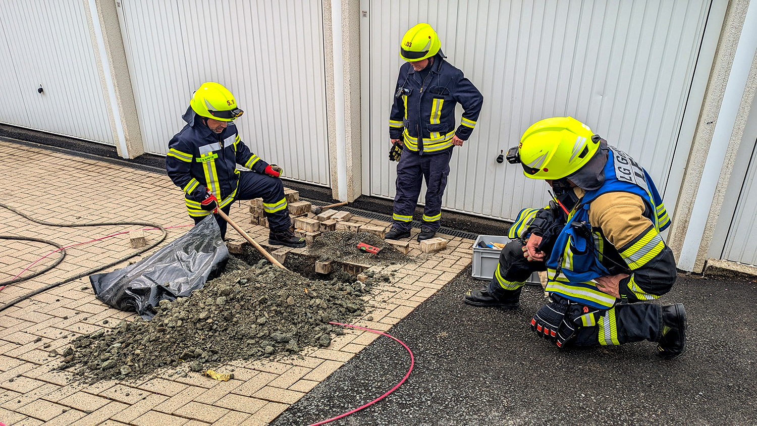 Drei Feuerwehrleute arbeiten an einem ausgehobenen Bodenloch vor weißen Garagentoren.