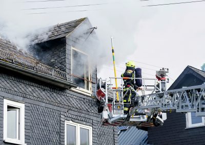 Ein Feuerwehrmann im Korb einer Drehleiter bekämpft einen Brand an einem schieferverkleideten Wohnhaus. Dichter Rauch dringt aus einem geöffneten Dachfenster und dem Dachstuhl.