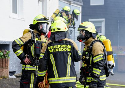 Drei Einsatzkräfte der Feuerwehr in Schutzkleidung mit Atemschutzmasken und gelben Sauerstoffflaschen besprechen sich. Im Hintergrund sind zwei weitere Feuerwehrleute an einer Haustür zu sehen.