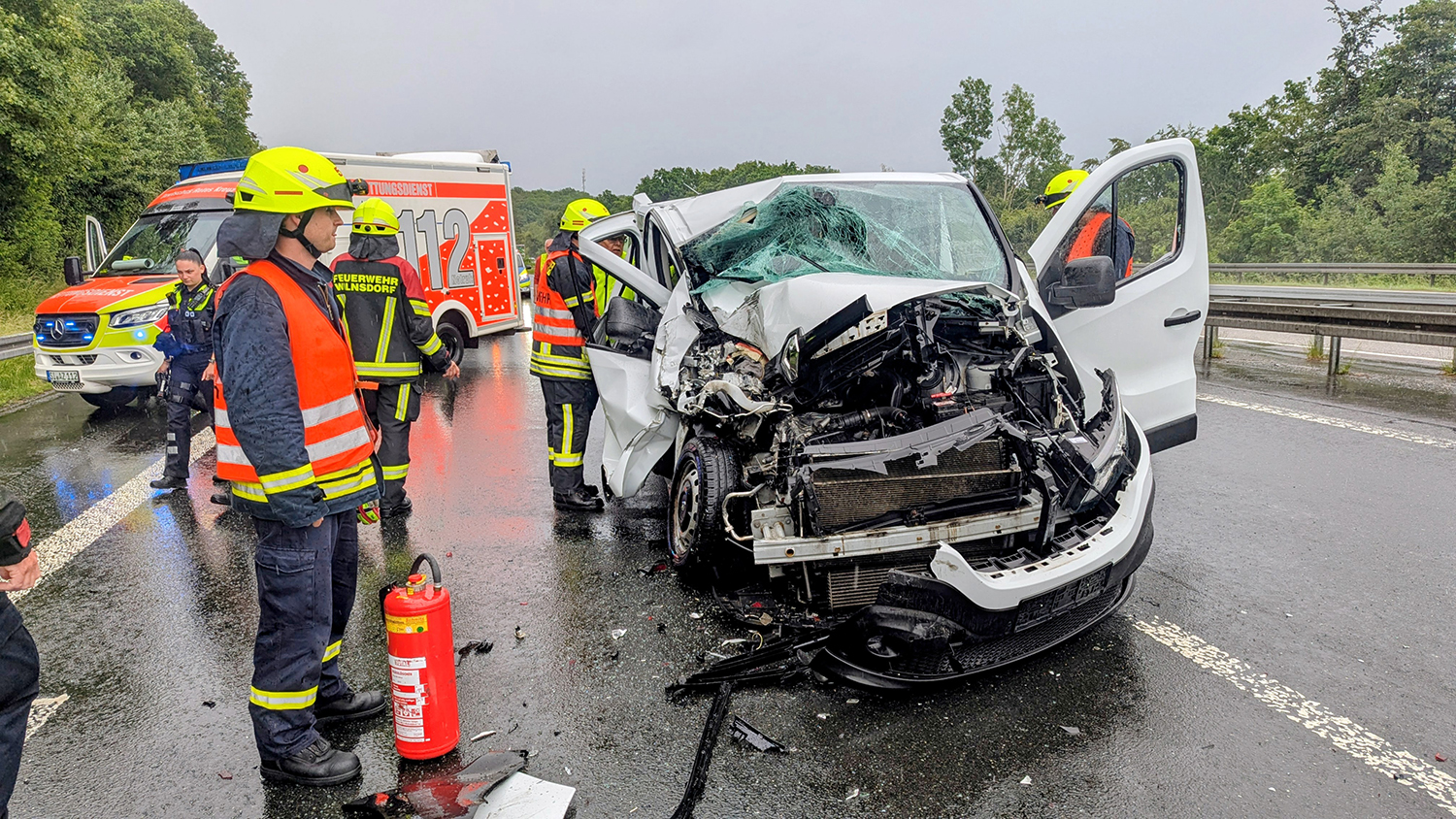 Kleinbus fährt auf Lastwagen – Sperrung der A45 Ein schwer beschädigter weißer Kleinbus nach einem Unfall auf einer nassen Autobahn. Kräfte von Feuerwehr und Polizei sind im Einsatz. Im Hintergrund ist ein Rettungswagen zu sehen.