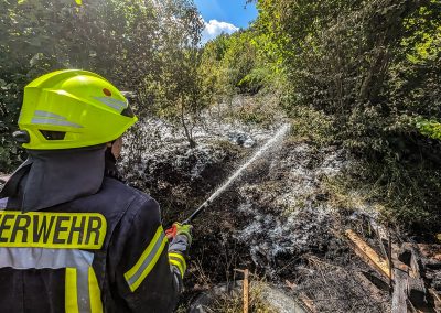 Feuerwehrkraft spritzt Wasser auf eine Brandstelle im Gebüsch.
