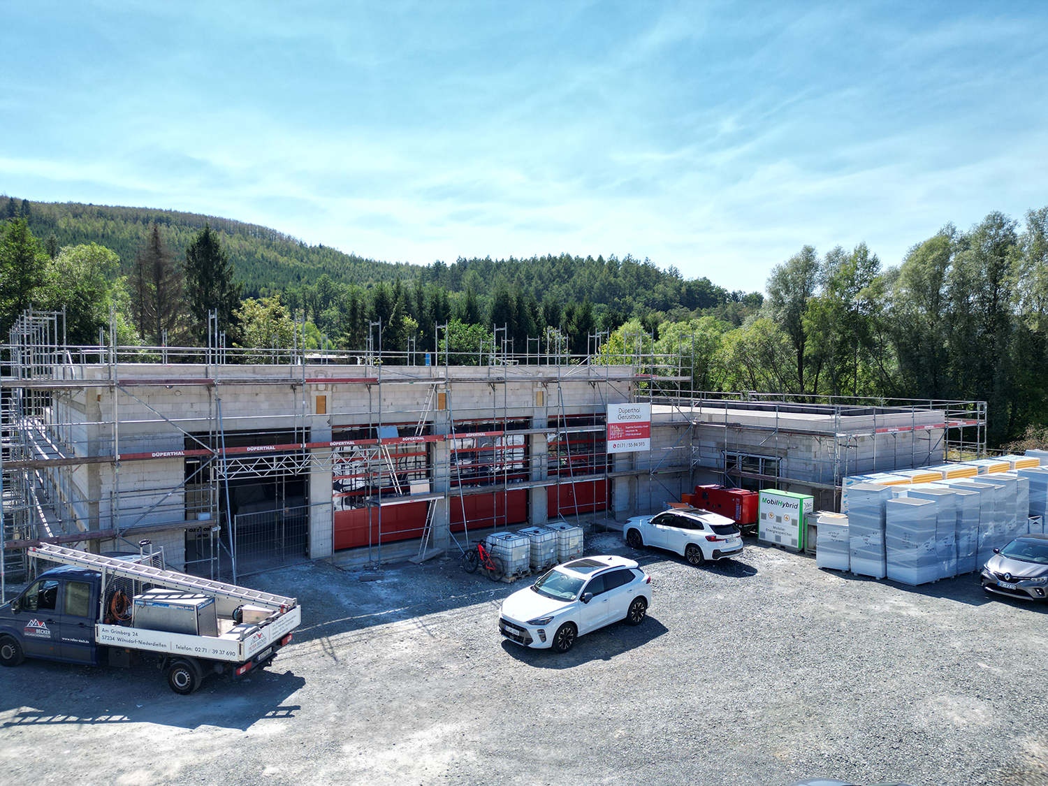 Außenaufnahme vom Neubau des Feuerwehrhauses im Rohbau mit Gerüst, davor geparkte Fahrzeuge und Baumaterialien.